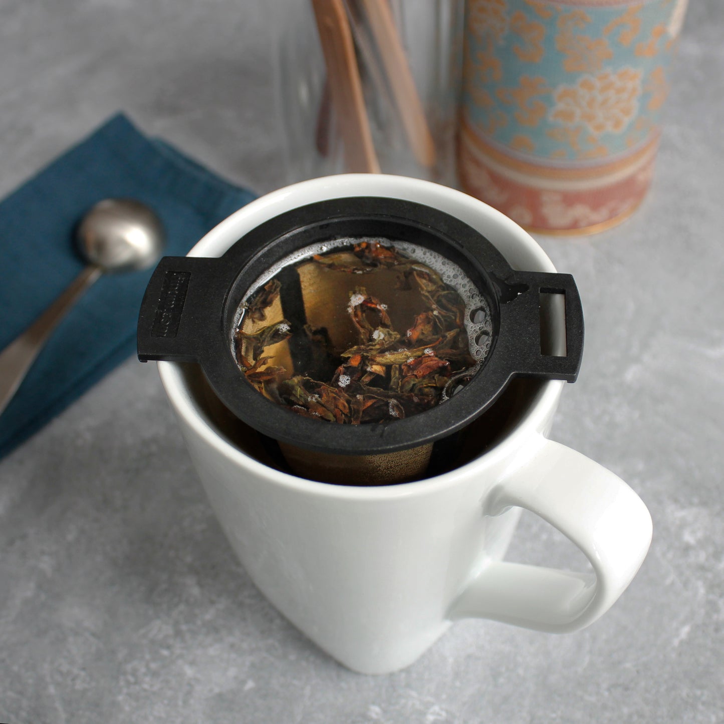 Black & Gold Strainer/Infuser - steeping in a white mug. a tea scoop can be seen in the background with other tea tools.