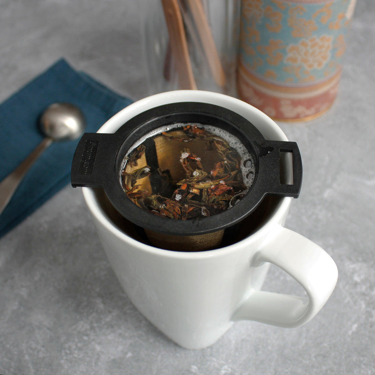 Black & Gold Strainer/Infuser - steeping in a white mug. a tea scoop can be seen in the background with other tea tools.