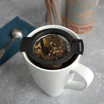 Black & Gold Strainer/Infuser - steeping in a white mug. a tea scoop can be seen in the background with other tea tools.