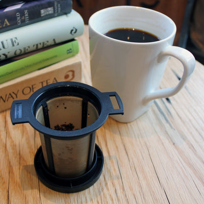 Black & Gold Strainer/Infuser - infuser sitting on it's lid. A cup of dark brewed tea in a white mug with books in the background on a table. 