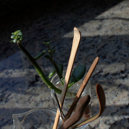 Bamboo Tea Rake - with other tools in a glass pitcher, a plant in a cup is behind it on a marble surface. 