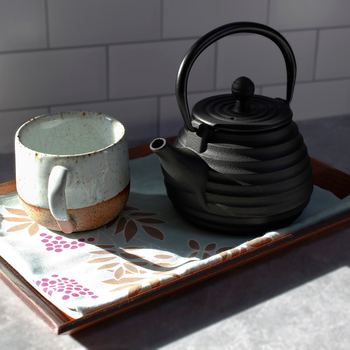 Black beehive teapot and ceramic speckled mug on a floral cloth with a white tiled wall background