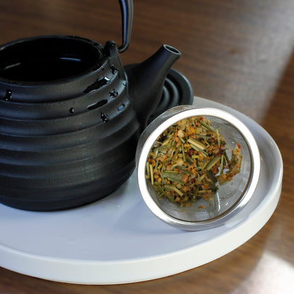 Black teapot with a metal tea infuser containing wet tea leaves on a white tray.