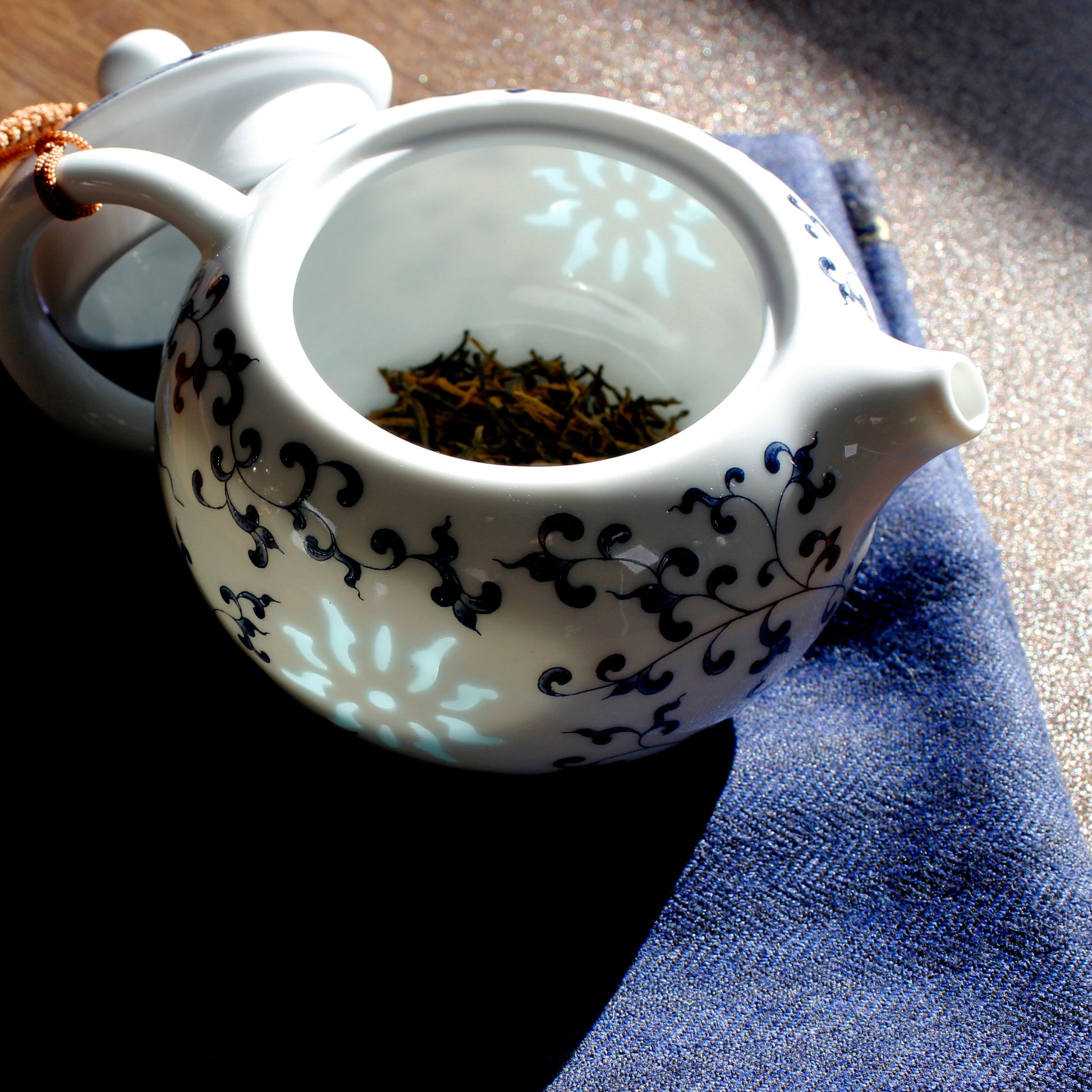 The teapot is open showing dried tea at the bottom. The light is glowing through the slightly transparent suns on the sides on the tea pot. The lid is leaning on the side and the whole thing is on a blue cloth. 