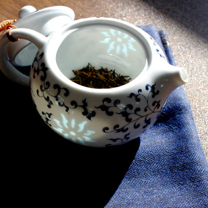 The teapot is open showing dried tea at the bottom. The light is glowing through the slightly transparent suns on the sides on the tea pot. The lid is leaning on the side and the whole thing is on a blue cloth. 