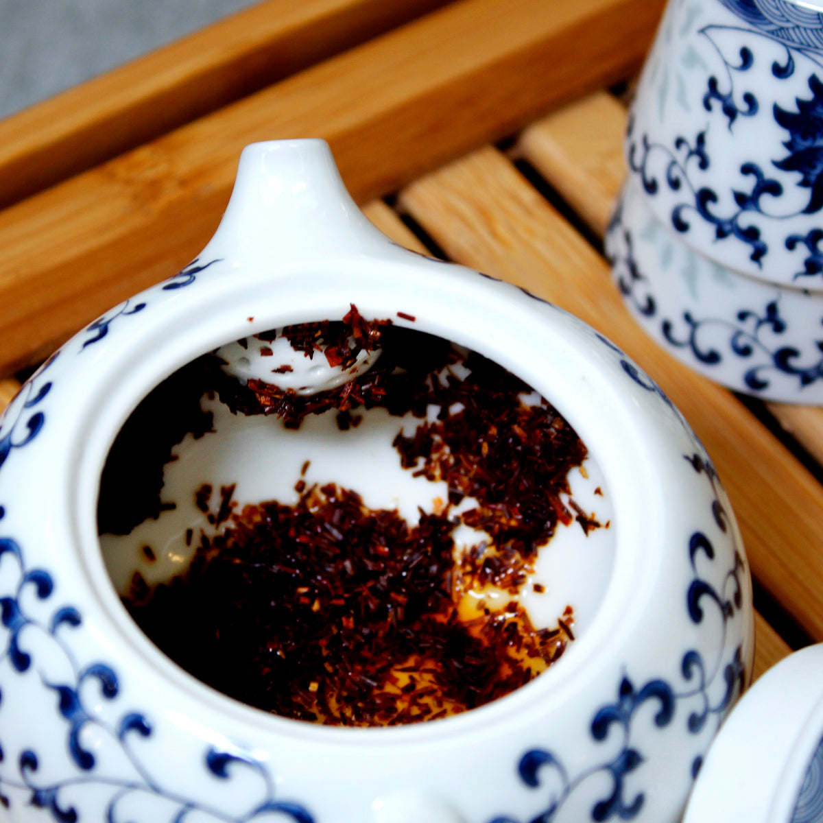 White teapot with blue flower and leaf patterns, wet tea leaves at the bottom with matching cups on the side. The set is on a wooden tray.