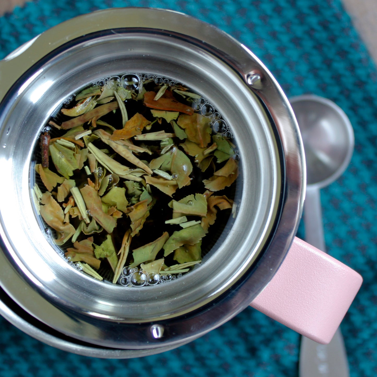 Tea leaves in the infuser floating in hot water, a scoop can be seen in the background on a blue and black patterned cloth.