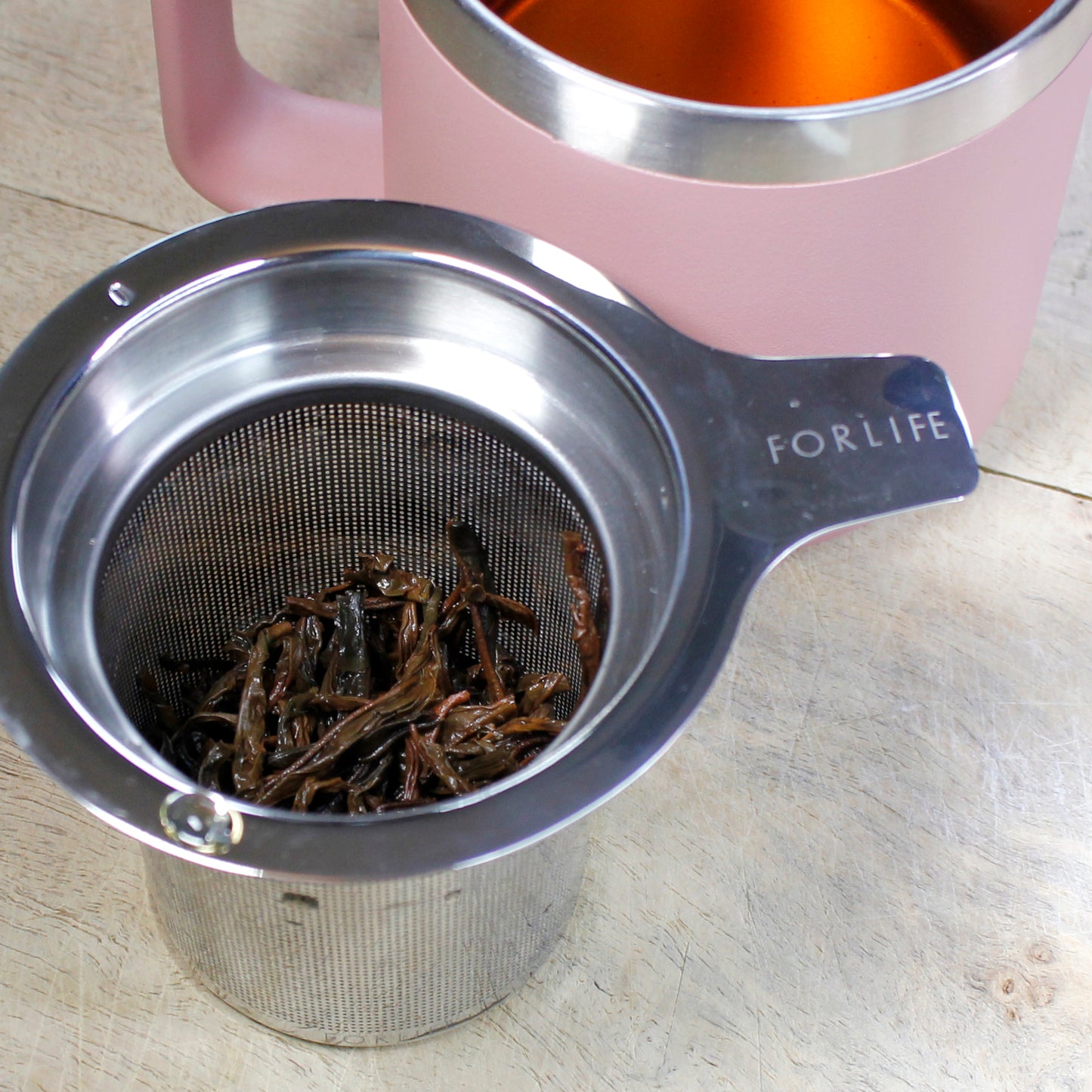Tea infuser with wet tea leaves next to a light pink mug on a light wood table. 