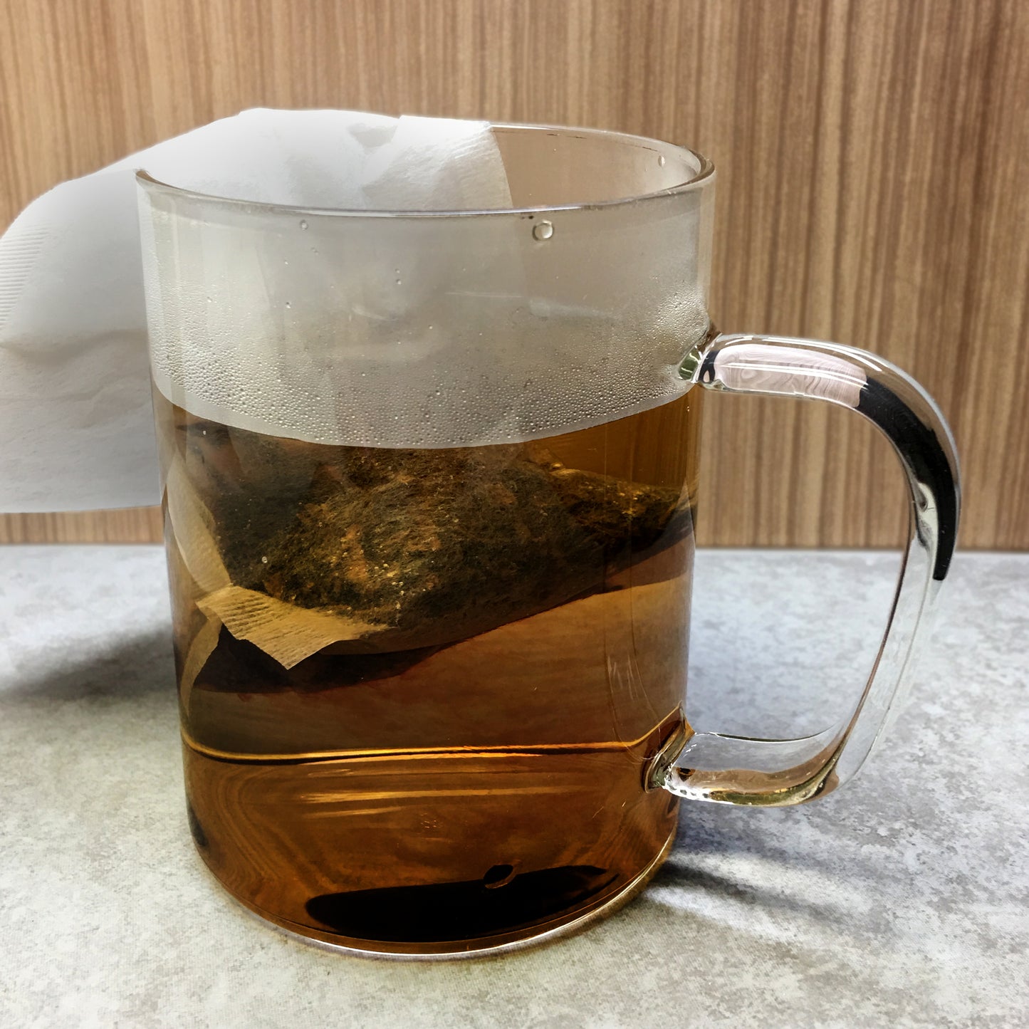 Tea bag in hot water in a glass pitcher on a grey table top, wood wall background. 