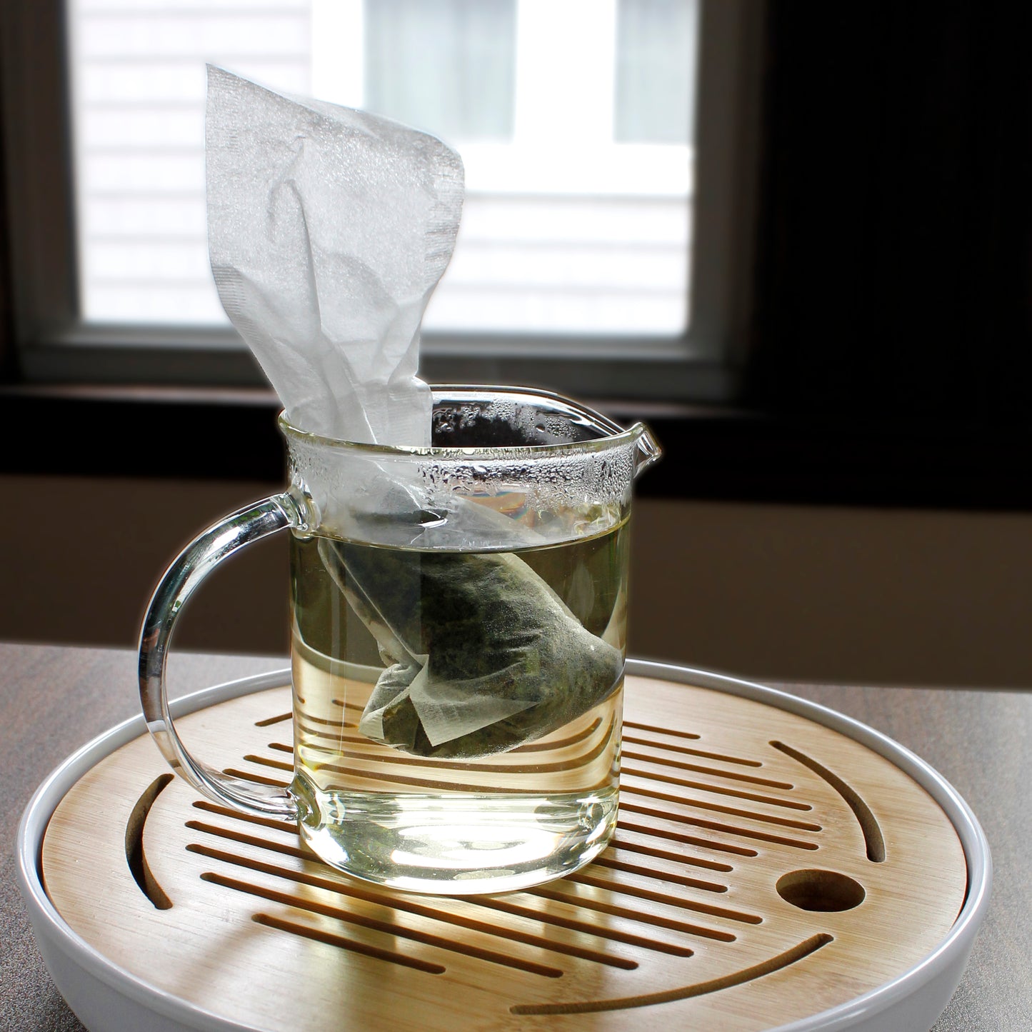 Fill-it-yourself tea bag in hot water being seeped in a glass pitcher on a bamboo tray near a window.  