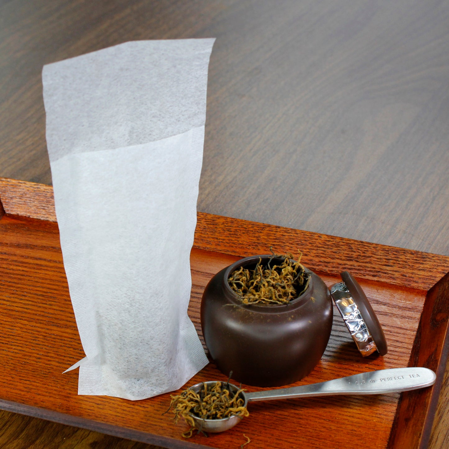 Fill-it-yourself tea bags with dried tea in a tea scoop and tea container next to the tea bag on a wood tray. 