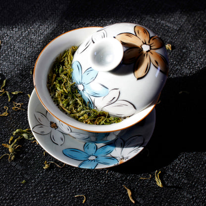 Ceramic gaiwan with floral design on a dark surface. The lid is tipped to show wet tea leaves, a few leaves have been scattered around the Gaiwan. 