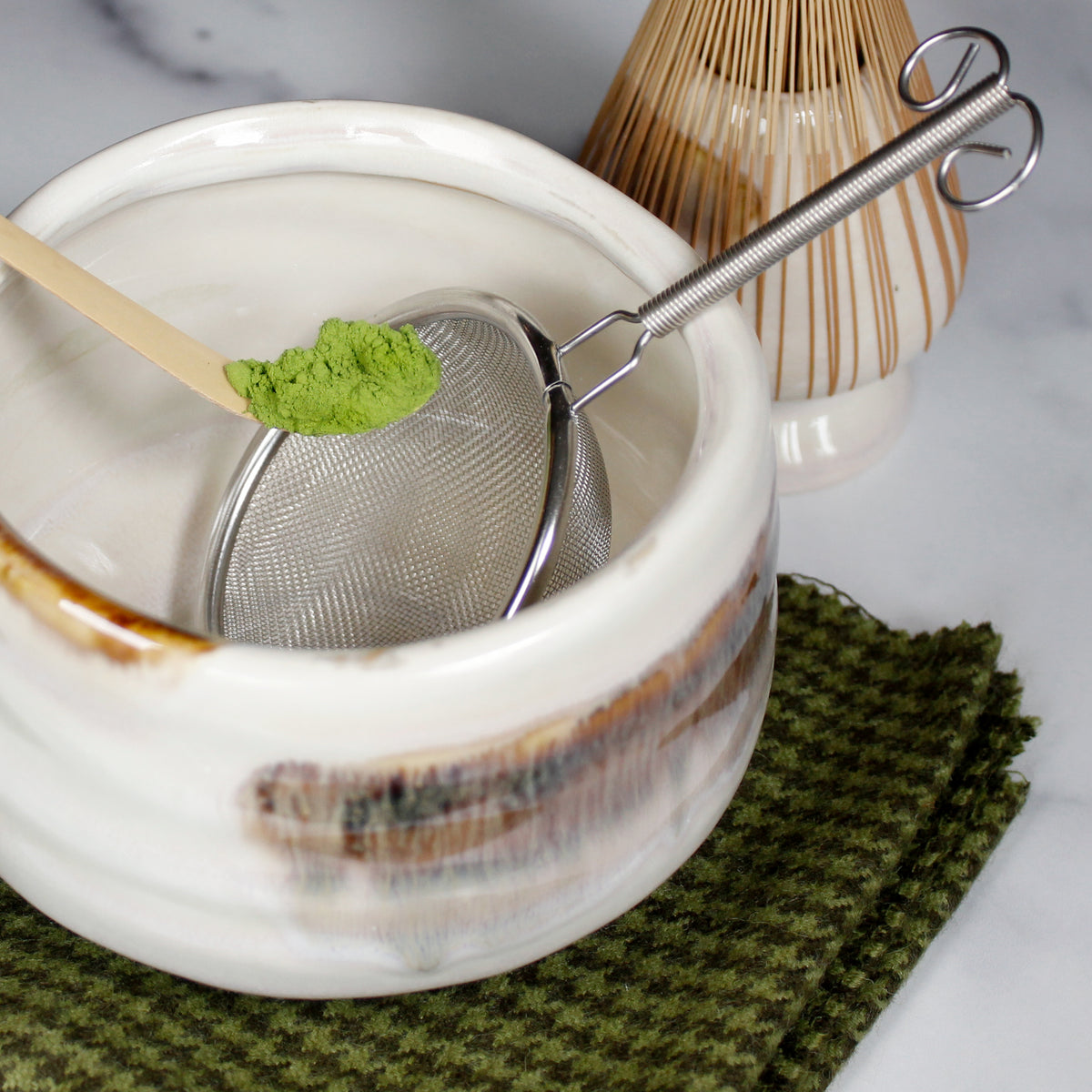Gabaron Ceremonial Grade Matcha - Marukyu Koyamaen - Uji, Kyoto, Japan - powder being spooned to a sifter with Matcha scoop into a white, brown and black stripped bowl sitting on a green cloth with a whisk in the background. Tasting Notes:
nori • wheat grass • unsweetened cocoa