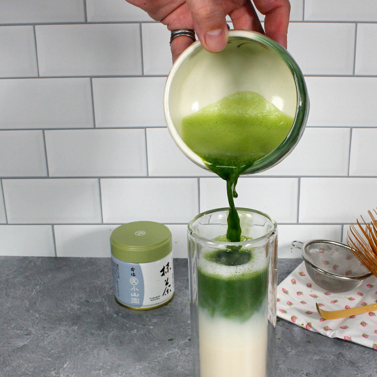 Gabaron Ceremonial Grade Matcha - Marukyu Koyamaen - Uji, Kyoto, Japan - latte, the matcha is being poured into a tall glass of milk. A tin, sifter, scoop and whisk are on the counter. Tasting Notes:
nori • wheat grass • unsweetened cocoa 