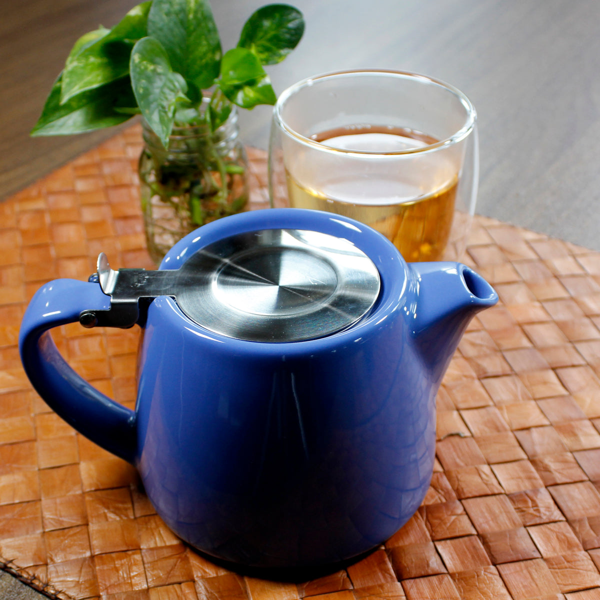 Blue teapot in front of a bubble glass cup and plant on a woven mat on a wood table. 