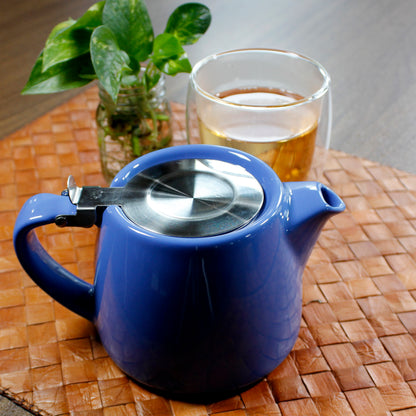 Blue teapot in front of a bubble glass cup and plant on a woven mat on a wood table. 