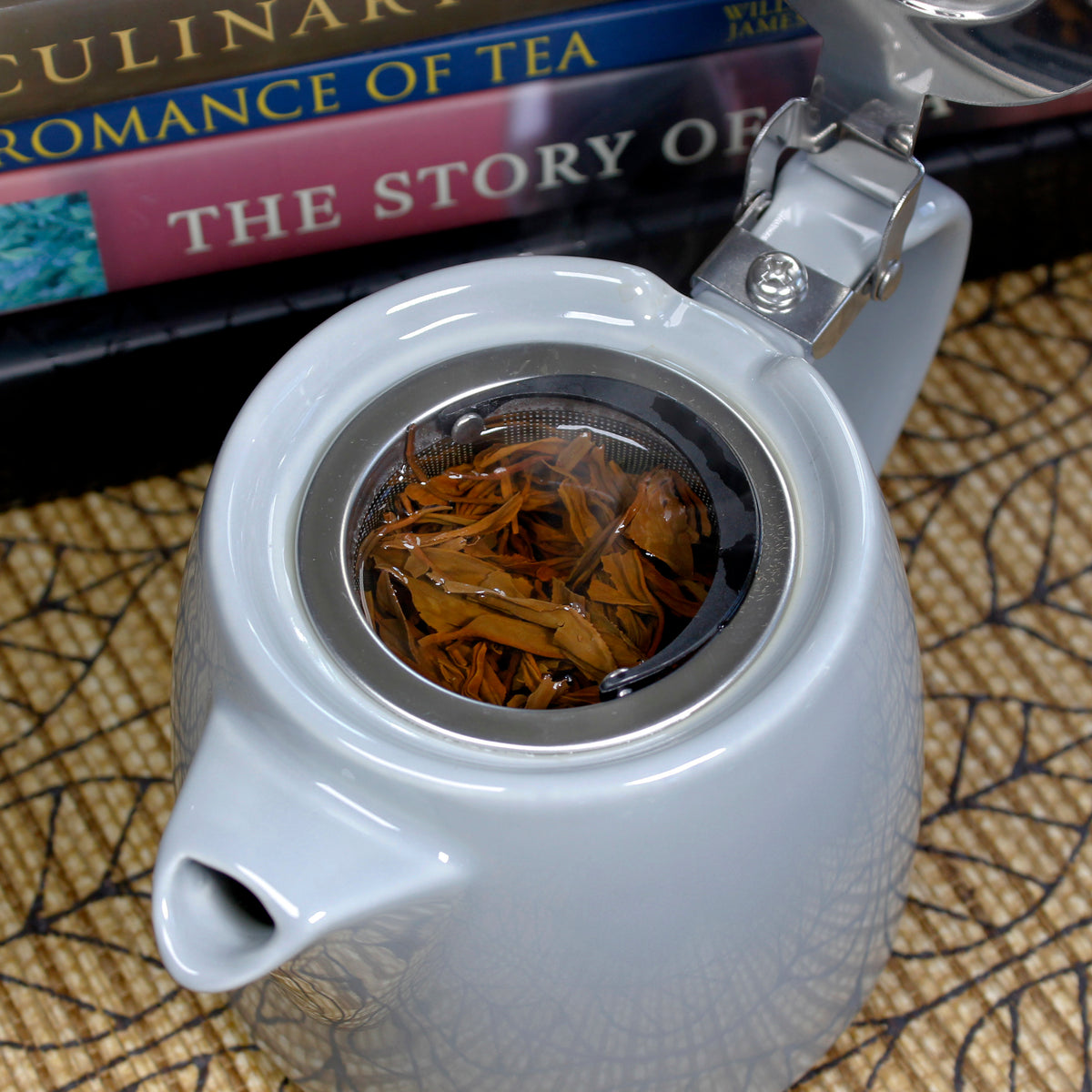 A ceramic teapot showing the inside with a strainer filled with tea. Books are in the background on a brown leaf printed cloth. 