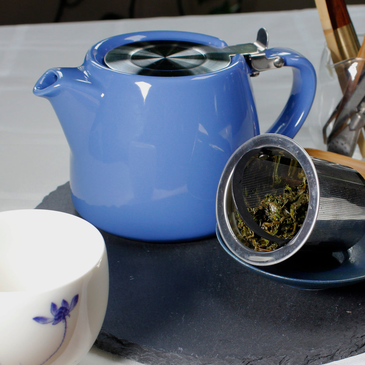 Blue ceramic flip lid teapot, the strainer on the side filled with wet tea leaves on a small dish and grey slate. A white tea cup with a small blue flower in the foreground.