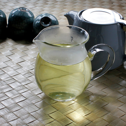 Dark grey ceramic teapot behind a glass pitcher filled with tea. Dark tea cups in the background on a woven table. 
