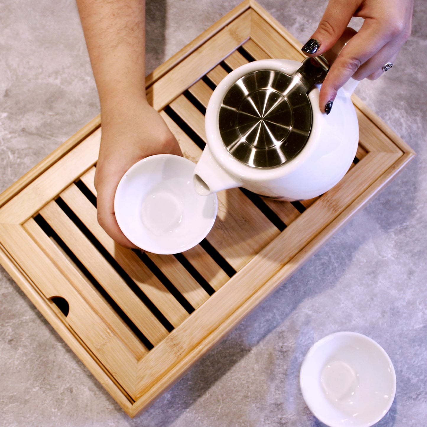 Person pouring tea from a white teapot into a white cup on a tray. A white cup is next to the bamboo tray. 
