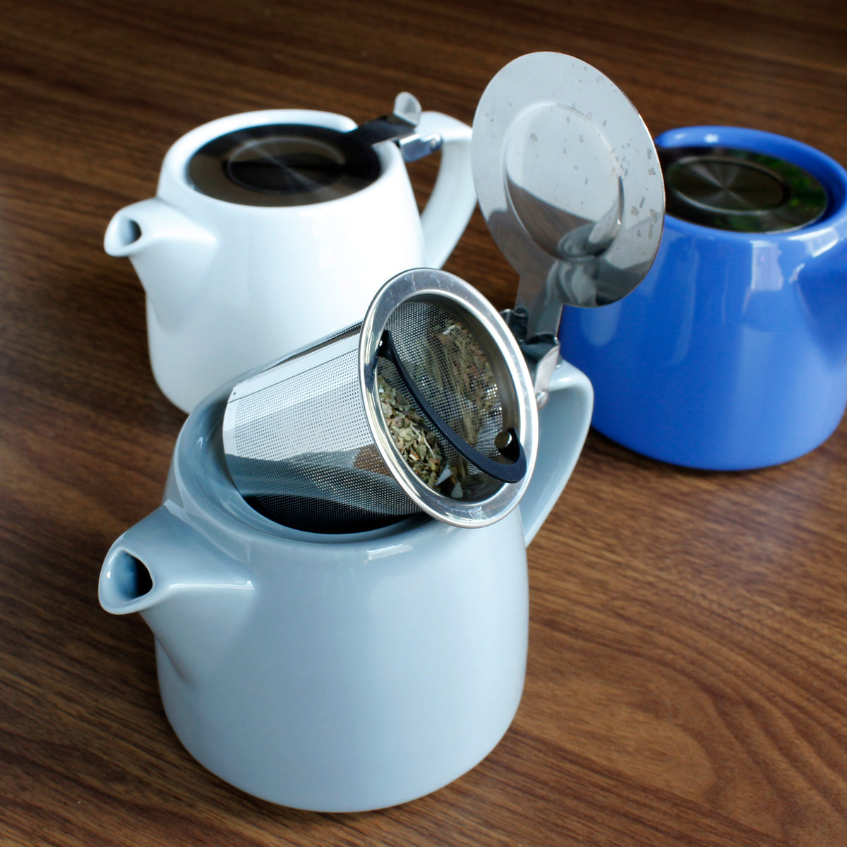 Three ceramic flip lid teapots, the colors are white, blue and light blue. One lid if open showing dried tea on a dark wood surface. 