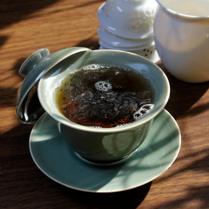 Green gaiwan with brewed tea in it, white stack of teacups and pitcher in background on a dark wood table. 