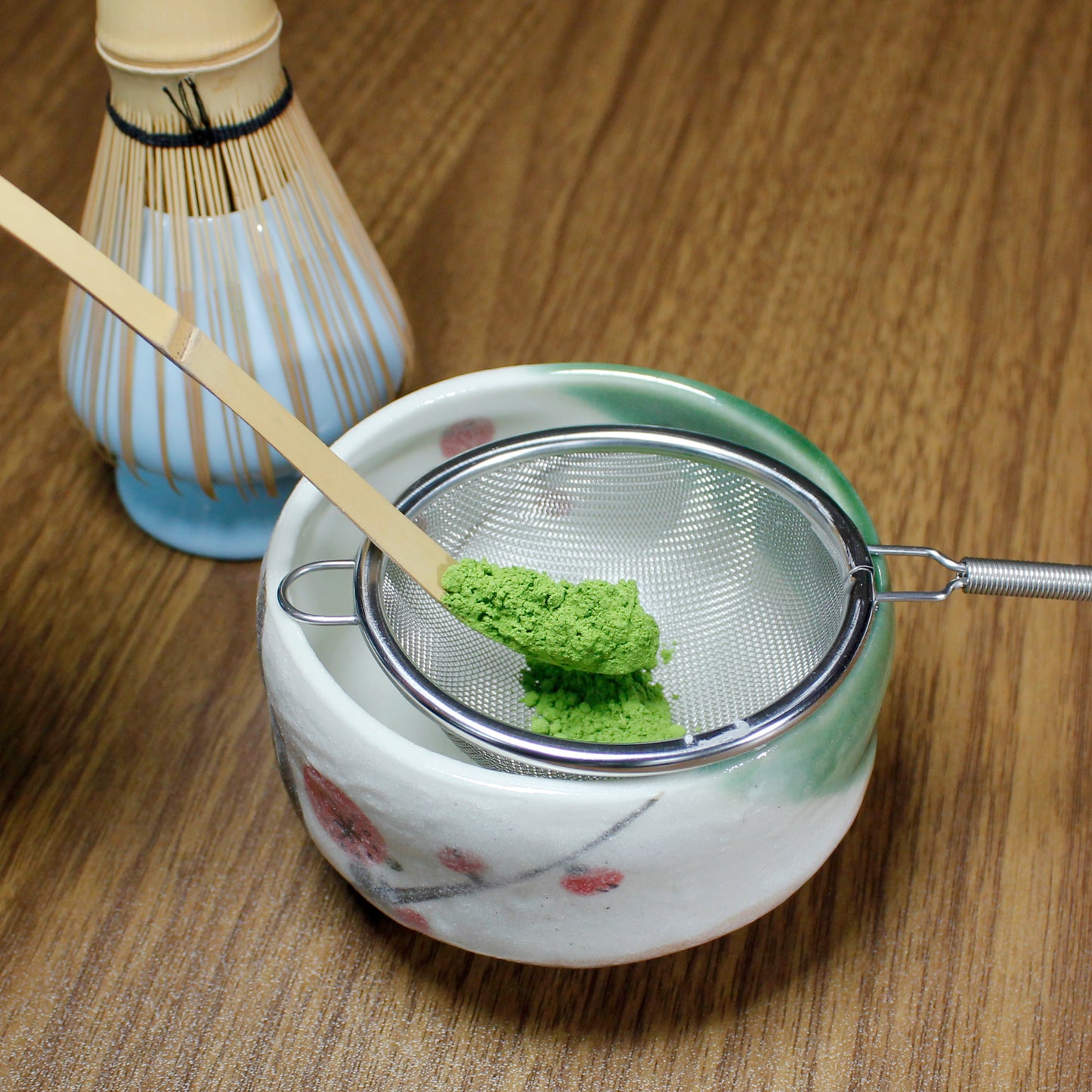 Matcha scoop holding matcha over a sifter resting on a matcha bowl. Behind is a bambo match whisk it its chasen holder. The whole set in on wood table.