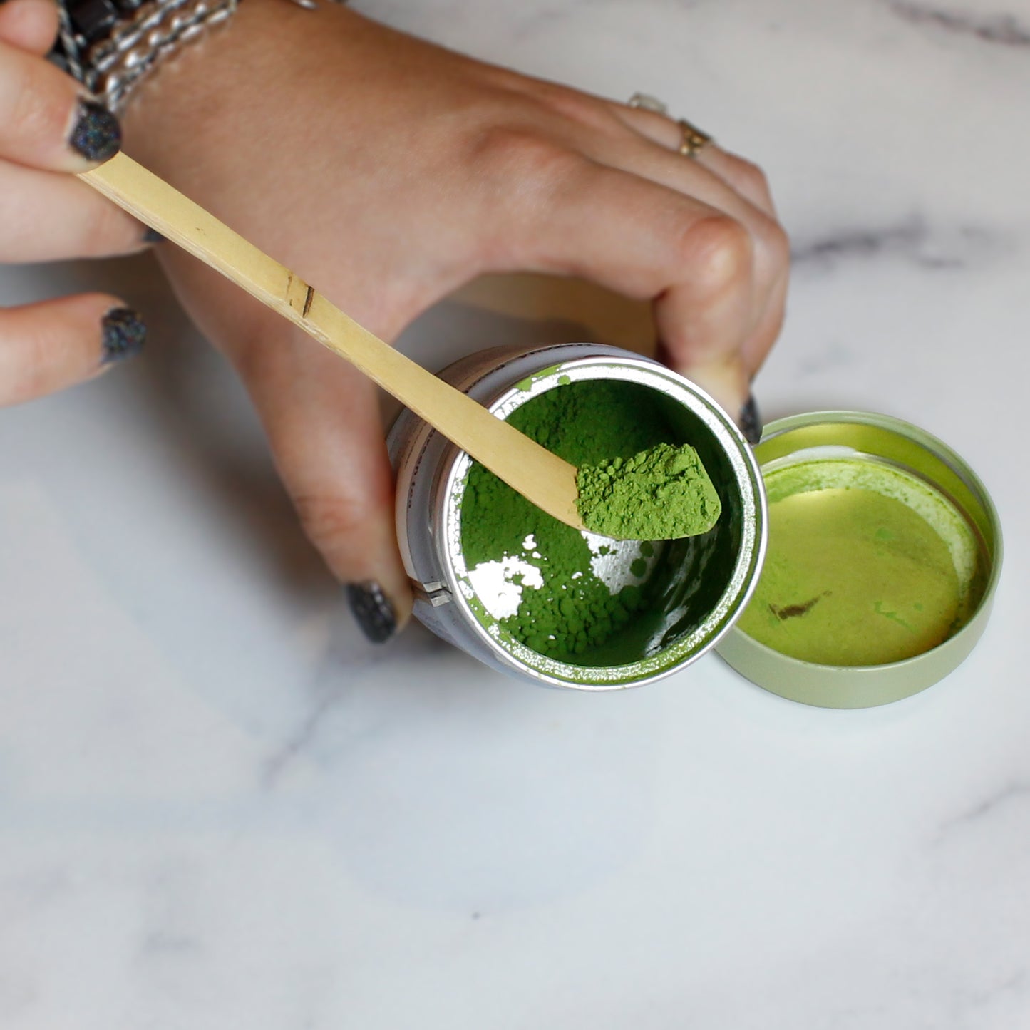 Person scooping green matcha powder from a container with a bamboo matcha scoop on a marble table. 