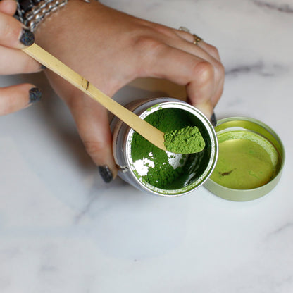 Person scooping green matcha powder from a container with a bamboo matcha scoop on a marble table. 
