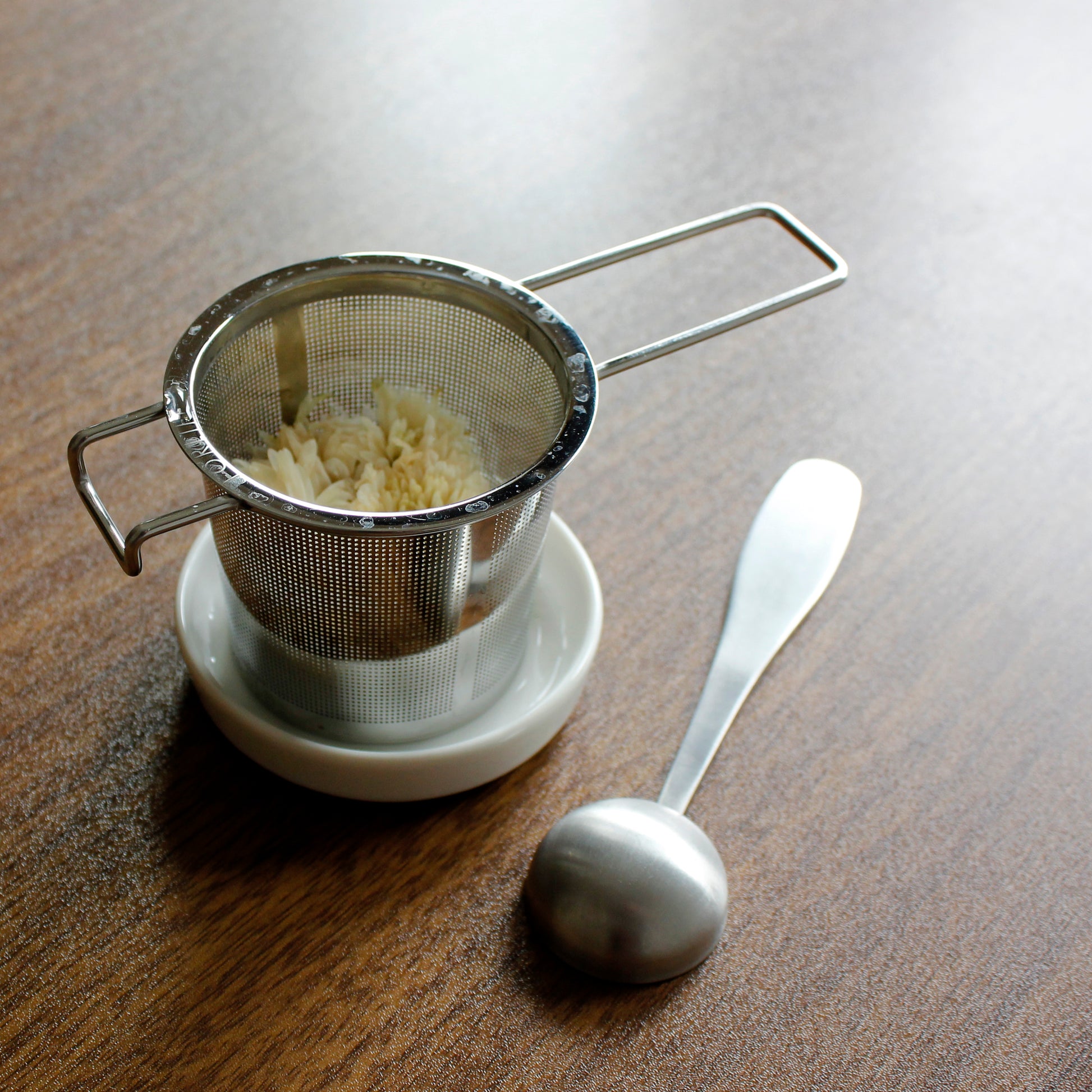 Strainer filled with wet tea on a resting dish with tea scoop next to it on a dark wood table.  