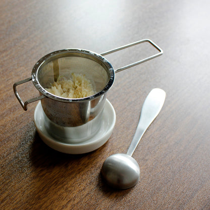 Strainer filled with wet tea on a resting dish with tea scoop next to it on a dark wood table.  
